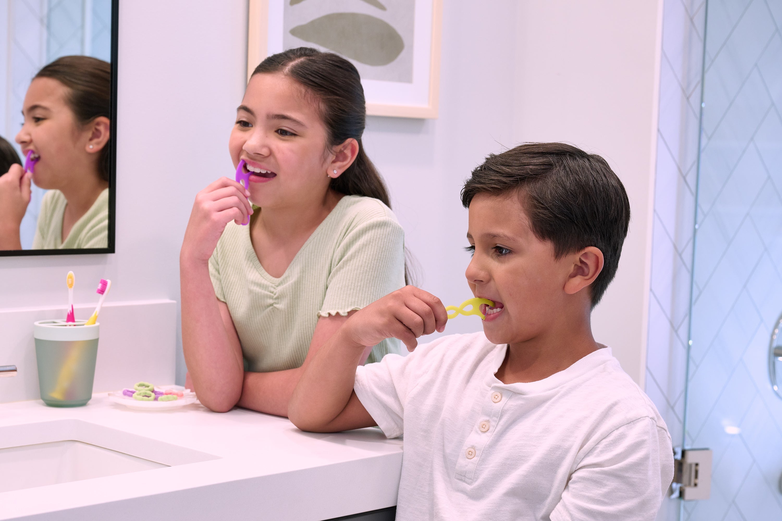 Two children flossing their teeth in a bathroom with a mirror reflecting their actions.